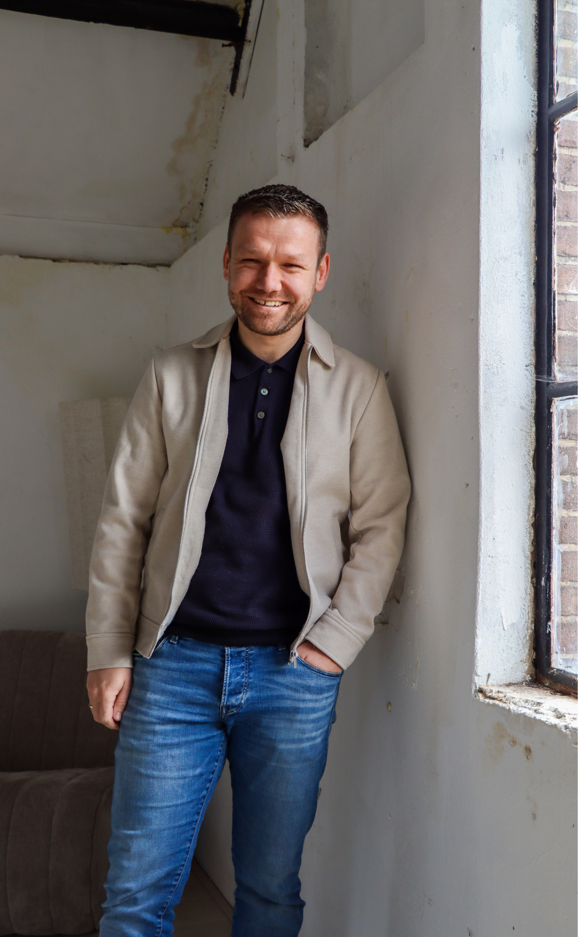 Smiling man in a beige jacket and dark polo leaning against a sunlit wall by a window.