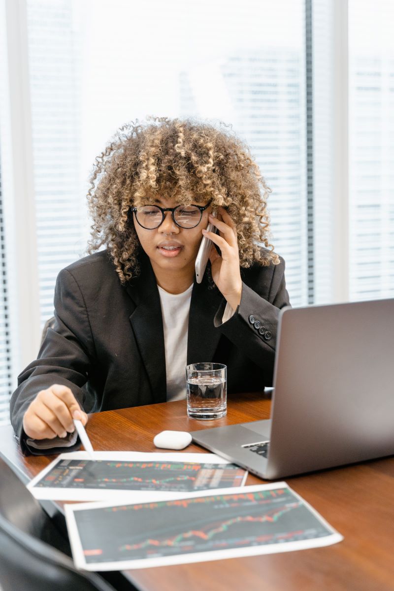 Een data analist analyseert de cijfers van een bedrijf en is tegelijk aan het bellen met voor haar een laptop en papier met daarop tabellen.